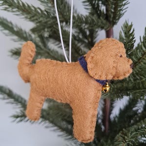 Könnte beinhalten: Handgefertigter Hundeanhänger aus Filz in Brauntönen, mit blauem Halsband und kleiner goldener Glocke. Der Hund hat ein einfaches, stilisiertes Design und wird an einer weißen Schnur aufgehängt, perfekt für die Weihnachtsdekoration.
