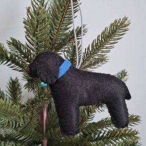 May include: Black felt dog ornament with a blue collar and a small bell hanging from the collar. The ornament is hanging from a white string on a green artificial Christmas tree.