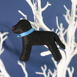 May include: Black felt dog ornament with a blue collar and a silver bell. The ornament is hanging from a white branch.