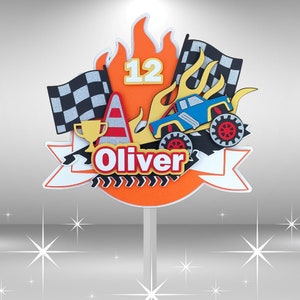 May include: Orange and yellow monster truck cake topper with a checkered flag, a trophy, a traffic cone, and the number 12. The topper also includes the name "Oliver" in yellow letters.