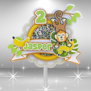 May include: A green, yellow, and brown cake topper with a monkey, bananas, and the name "Jasper" and the number "2".