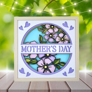 Puede incluir: Un marco de madera con un diseño de papel en capas de flores en morado y azul y el texto "MOTHER'S DAY".