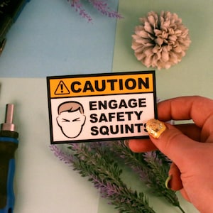 May include: A rectangular sticker with a yellow and black border, featuring the word "CAUTION" above the phrase "ENGAGE SAFETY SQUINTS." The sticker includes a graphic of a person squinting. The sticker is held by a hand with gold glitter nail polish.