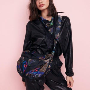 Puede incluir: Un bolso bandolera negro con un colorido estampado geométrico. El bolso se lleva sobre un atuendo negro, y la correa presenta un diseño similar. El fondo es de color rosa liso.
