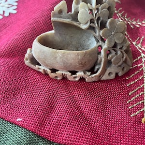 May include: A carved stone bowl with floral and leaf details. The bowl has a small, rounded basin and is supported by a base with intricate carvings. The color is a light beige, and the bowl is set against a red fabric background.