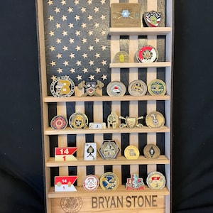 May include: Wooden display case in the shape of the American flag, with a black background and wooden stars. The case has multiple shelves holding various military challenge coins and insignia. The name "BRYAN STONE" is engraved at the bottom.