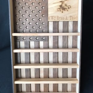 May include: A rectangular wooden display shelf featuring a vertical American flag design with alternating dark and light wood stripes. The upper left corner has cut-out stars, and the upper right section displays an engraved helicopter and the text CW2 Ryan A. Thiel.