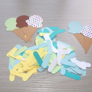 May include: A collection of paper cutouts featuring ice cream cones in light green, brown, and blue, alongside a pile of small, rectangular paper strips in shades of yellow, light green, white, turquoise, and light blue for crafting projects.