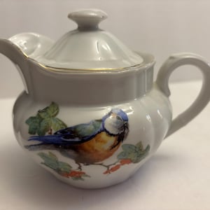 May include: White ceramic creamer with a lid, featuring a painted blue, orange, and brown bird with green leaves and red berries. The creamer has a gold trim and a curved handle. A decorative piece for serving cream or other liquids.