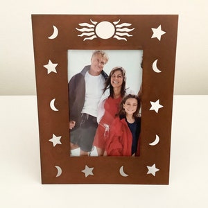 May include: A brown wooden picture frame with a white sun and stars design. The frame has a cutout design of moons and stars around the edge. The frame is holding a photo of a family.