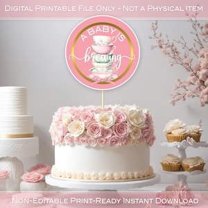 以下が含まれることがあります： 「A Baby Is Brewing」というテキストと積み重ねられたティーカップが描かれたピンクとゴールドの印刷可能なケーキトッパー。