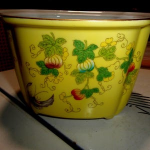 May include: A bright yellow bowl with a scalloped rim and a gold-tone edge. The exterior features a repeating pattern of green leaves, curling vines, and small orange and blue pumpkins. The bowl has a smooth, glossy finish and a wide opening.
