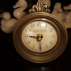 May include: A vintage brass clock with a cream-colored face and black hands. The clock face features the words "MADAME GISELLE" and Roman numerals. The clock is surrounded by a decorative brass frame and is set against a dark background.