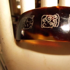 May include: A wide, dark brown, translucent bangle bracelet featuring a tortoiseshell pattern. The surface is decorated with a series of engraved, light-colored, square-shaped glyphs or symbols that wrap around the exterior of the circular band.