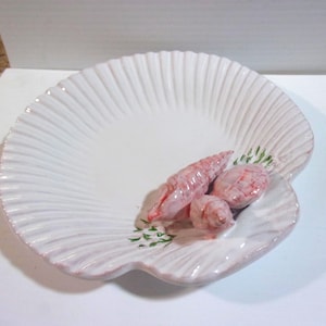 May include: A white ceramic dish shaped like a large scallop shell, featuring a ribbed texture and a glossy finish. Three small, pink, textured seashell sculptures are attached to the surface, accented with small green painted leaf details.