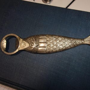 May include: A silver-toned metal bottle opener shaped like a fish. The design features detailed scales along the body, a defined head with eye markings, and a flared tail fin. The circular opening for the bottle cap is integrated into the fish head.
