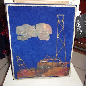 May include: A blue velvet framed wall art piece with a copper metal sculpture of a ship with a tall mast and a smaller mast. The sculpture is on a blue background with the text "SAFAVIEH CARE" in white.