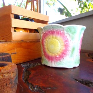 May include: A ceramic planter featuring a raised, embossed sunflower design with a bright yellow center and pink petals, set against a pale green background. The planter has a scalloped rim and a textured surface with a glossy finish.