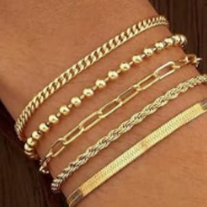 May include: A set of five gold-toned bracelets worn together. The collection includes a curb chain, a beaded chain, a rectangular link chain, a twisted rope chain, and a flat herringbone chain, all featuring a polished metallic finish.