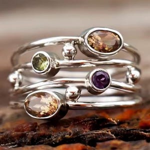 May include: A multi-band silver ring featuring four distinct gemstones set in individual bezels. The ring includes two oval-cut brown stones, one round-cut green stone, and one round-cut purple stone, accented by small silver spheres between the bands.