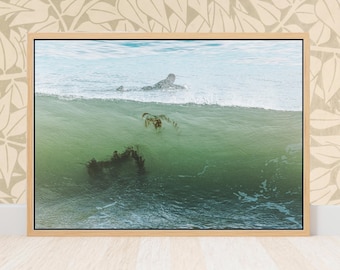 Può includere: Fotografia incorniciata di un surfista nell'oceano. L'immagine cattura un momento in cui il surfista pagaia attraverso un'onda. L'acqua è un mix di turchese e verde, con alghe visibili sotto la superficie. La cornice è di colore legno chiaro.