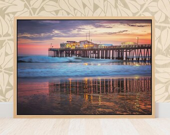 May include: A framed photograph of a pier extending into the ocean at sunset. The sky is a mix of pink, orange, and purple hues. The pier's lights reflect in the wet sand, creating a symmetrical image.