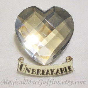 May include: A clear crystal heart-shaped charm with a silver finish and the word "UNBREAKABLE" printed on a scroll banner below.