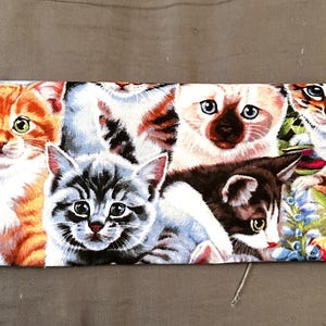 May include: A fabric panel featuring a collage of adorable cat faces. The cats have various fur patterns, including orange tabby, Siamese, and calico. Some cats are surrounded by flowers, with a gray background.