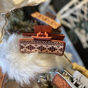 May include: A terracotta-colored hair clip with a geometric pattern. The clip has a spring-loaded mechanism and is designed to hold hair in place.