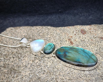 Puede incluir: Un collar de plata con un colgante que presenta tres piedras: una labradorita azul grande de forma ovalada, una labradorita azul redonda más pequeña y una piedra de luna blanca.