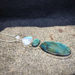 May include: A silver necklace with a pendant featuring three stones: a large oval-shaped blue labradorite, a smaller round blue labradorite, and a white moonstone.