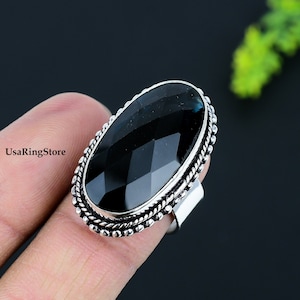 Puede incluir: Un anillo de plata con una gran piedra negra facetada engastada en un bisel ovalado decorativo. El anillo se lleva en un dedo.