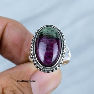 May include: Oval ring with a polished, deep red and green gemstone set in a silver-coloured metal band. The band has a textured, rope-like border. The ring is held by a hand. The gemstone is the focal point.