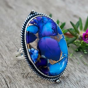 Puede incluir: Un anillo de plata con una gran piedra ovalada que presenta un vibrante patrón de mosaico con vetas de color púrpura intenso, azul brillante y cobre metálico. La piedra está engastada en un borde de plata con diseño de cuerda trenzada.
