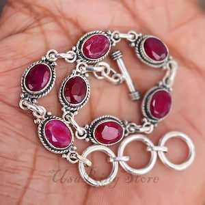May include: A silver bracelet featuring oval-shaped, deep red gemstones. The gemstones are set in a decorative silver frame, connected by silver links. The bracelet has a toggle clasp and is displayed on a hand.