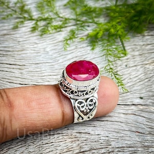 Puede incluir: Un anillo de plata con un diseño en forma de corazón y una gran piedra de rubí ovalada. El anillo se lleva en un dedo.