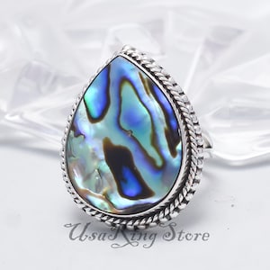 May include: A pear-shaped ring featuring a vibrant abalone shell with iridescent blues, greens, and purples. The shell is set in a silver-toned metal with a braided rope design around the edge. The text "UsaKing Store" is visible.