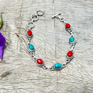 May include: A silver chain bracelet featuring alternating oval-shaped red and turquoise-coloured stones set in decorative metal bezels. The bracelet includes a toggle clasp closure with a series of circular links at one end for adjustable sizing.