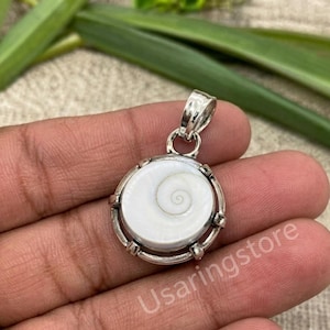 Puede incluir: Un colgante circular que presenta una concha de ojo de Shiva blanca con un patrón de espiral natural, engastada en un marco de metal plateado pulido con una reasa decorativa. La pieza incluye una pequeña anilla metálica en la parte superior.