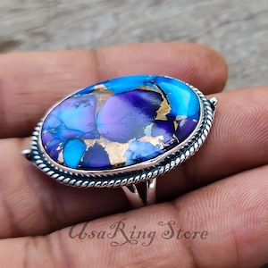 May include: Oval-shaped ring featuring a vibrant, multi-colored stone with shades of blue, purple, and gold. The stone is set in a silver-toned metal band with a textured, rope-like border. The ring is a piece of jewelry.