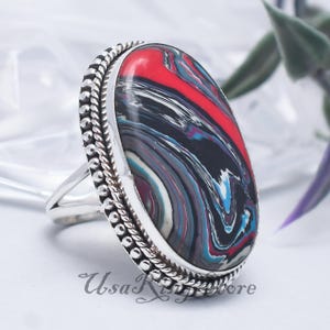 May include: Oval ring featuring a large, patterned stone in red, black, blue, and white. The stone is set in a silver band with a textured edge. A striking piece of jewellery.
