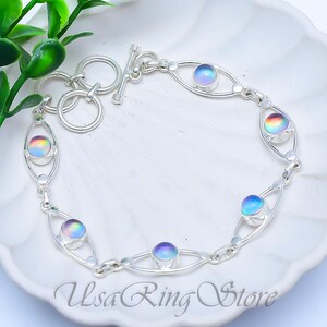 May include: A silver bracelet featuring oval links with iridescent, round gemstones. The bracelet has a toggle clasp and is displayed on a white surface. The text "Usa Ring Store" is visible.