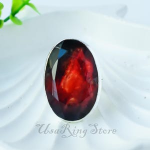 Pode incluir: Um anel de prata com uma grande pedra vermelha de forma oval. A pedra preciosa tem um fundo escuro, quase preto, com um padrão vermelho, laranja e amarelo.