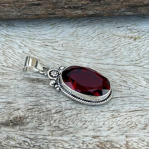 Può includere: Un ciondolo con una grande pietra preziosa rosso scuro dal taglio ovale, incastonata in una montatura in argento lucido. La montatura presenta dettagli metallici lavorati a perline e un piccolo motivo floreale sulla parte superiore.
