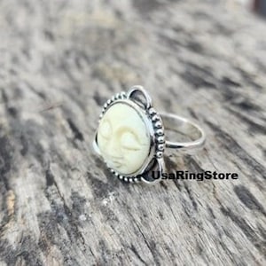 May include: A silver ring featuring a white bone carving of a face with closed eyes. The ring is set in a silver bezel with a beaded edge.
