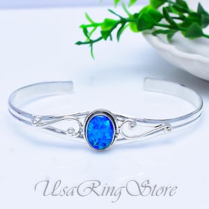 May include: A silver cuff bracelet with an oval, blue opal centre stone. The bracelet has decorative swirls either side of the opal. The text "Usa Ring Store" is at the bottom of the image.