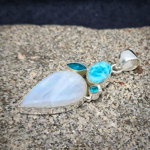 Könnte beinhalten: Ein silberner Anhänger mit einem tropfenförmigen Mondstein, umgeben von drei kleineren blauen Steinen.