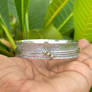 May include: A silver bangle bracelet with a textured surface and three gold beads. The bracelet is open and has a wide band.