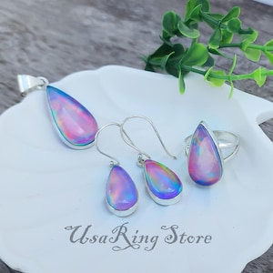 Peut inclure: Un ensemble de bijoux comprenant un pendentif, des boucles d'oreilles et une bague. Chaque pièce présente une pierre d'opale irisée en forme de goutte d'eau sertie dans un métal argenté. Les pierres d'opale affichent des teintes roses, violettes et bleues. L'ensemble est présenté sur une surface blanche.