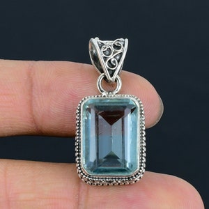 Puede incluir: Un colgante de plata con una piedra preciosa de aguamarina azul claro. El colgante tiene un diseño de filigrana y está engastado en una forma cuadrada.